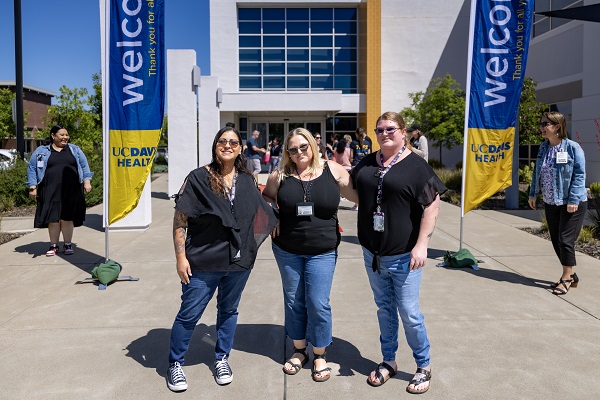 Three attendees stand smiling between two vertical UC Davis Health &ldquo;Welcome&rdquo; banners outside a building entrance.