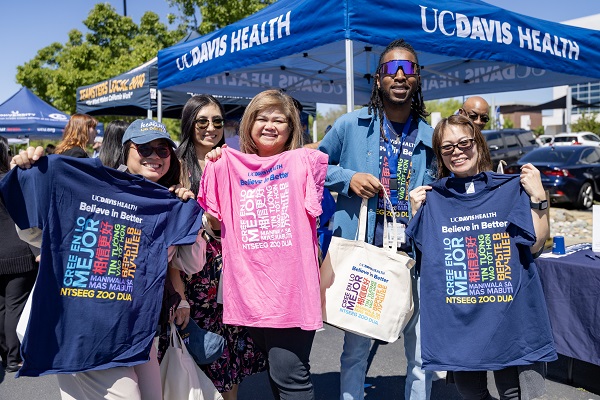 Five attendees stand together holding UC Davis Health &ldquo;Believe in Better&rdquo; T‑shirts in blue and pink at an outdoor event booth.