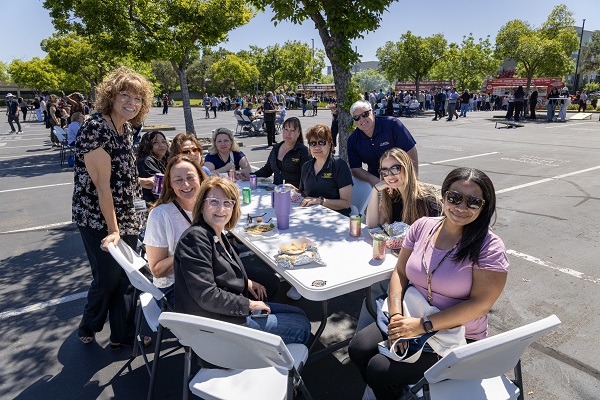A group of attendees sits and stands around a table eating food at an outdoor event.