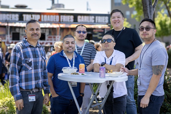Six attendees stand around a tall table with food and drinks, with a food truck in the background.