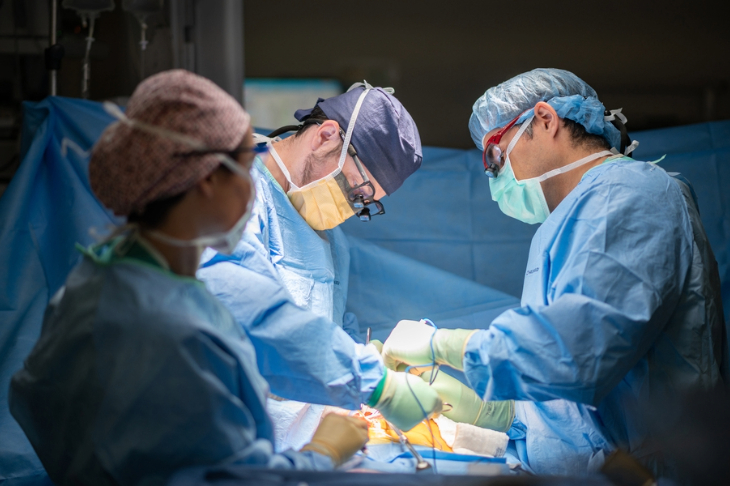 Three medical professionals in surgical gowns and masks perform a surgery under bright lights in an operating room.