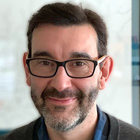 Smiling man with glasses and short dark hair, in a head-and-shoulders portrait against a light background.