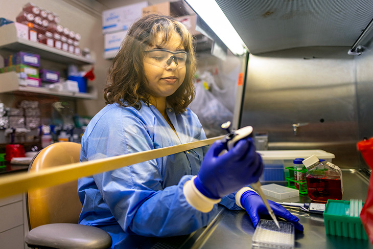 A person in a blue lab coat, gloves and protective eyewear working in a laboratory.