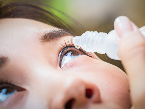 A woman holds an eye drop vial with a single drop above her eyes against a blurred background. 