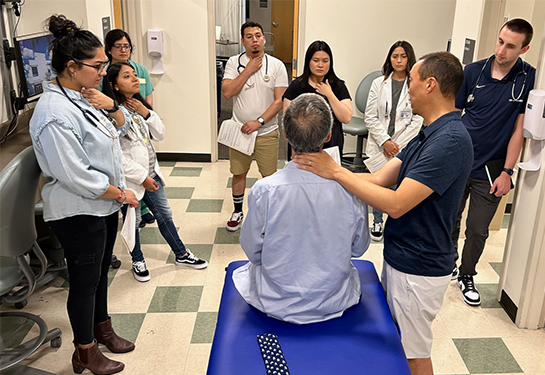 A group of medical students watch two professors demonstrate at a portable exam table how to check a neck for abnormalities.