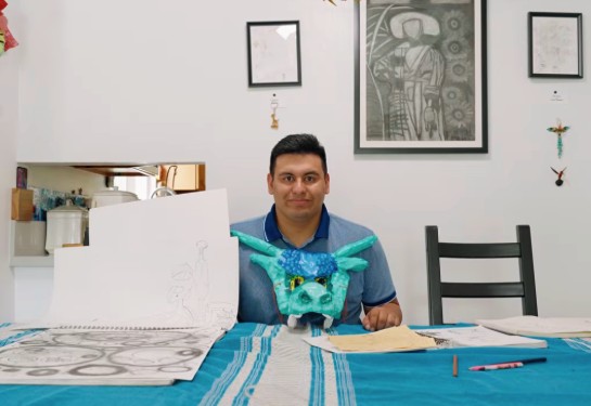Man seated at a table displaying artwork, including a hand-drawn sketch on paper and a colorful sculpted mask, with framed art hanging behind him