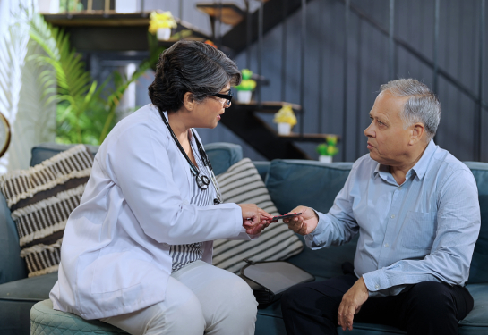 Female doctor visiting senior male patient in house and sitting on chair giving him medicine tablets.