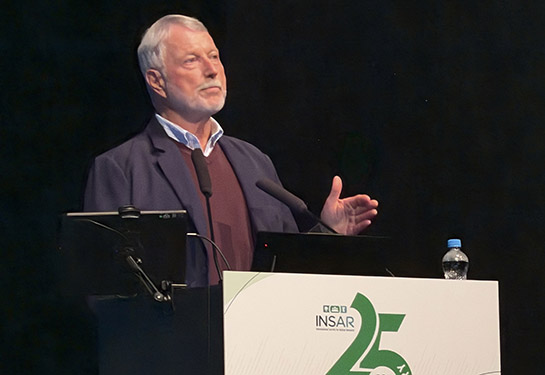 David Amaral, in a dark suit, stands behind a podium on the stage at the INSAR conference after accepting his Lifetime Achievement Award. 