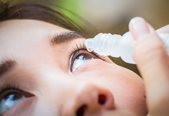 A woman holds an eye drop vial with a single drop above her eyes against a blurred background. 