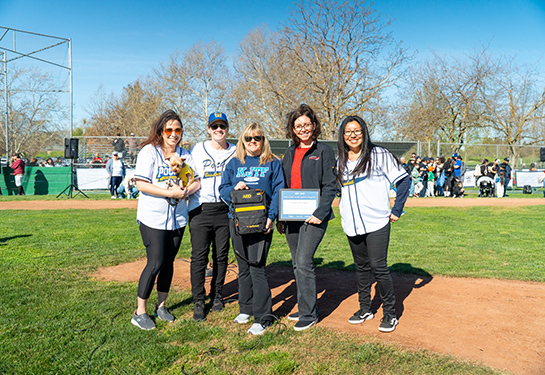 Five people stand on baseball field, with one person holding a certificate and one person holding an AED in a bag.  