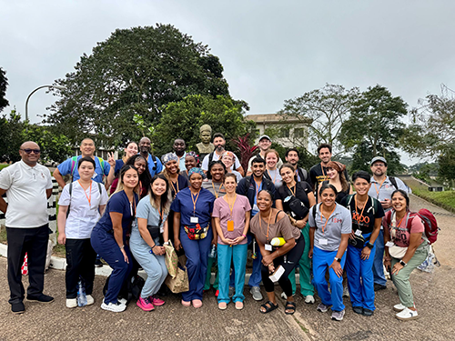 A large group of healthcare professionals wearing scrubs and badges pose outdoors, with trees and buildings in the background.