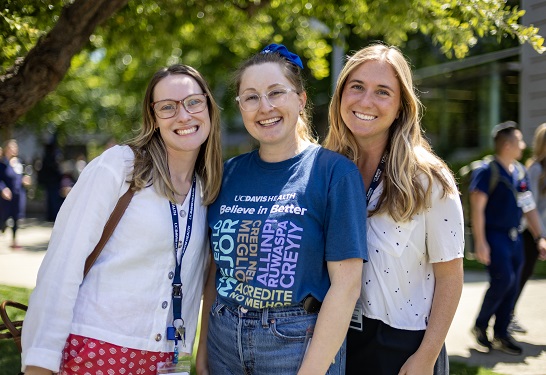 Three employees stand together outdoors wearing UC Davis Health apparel, posing for a photo on campus.