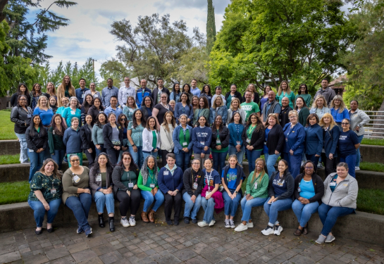 Dozens of people pose together outdoors in four rows on tiered stone seating, with trees and campus greenery in the background.