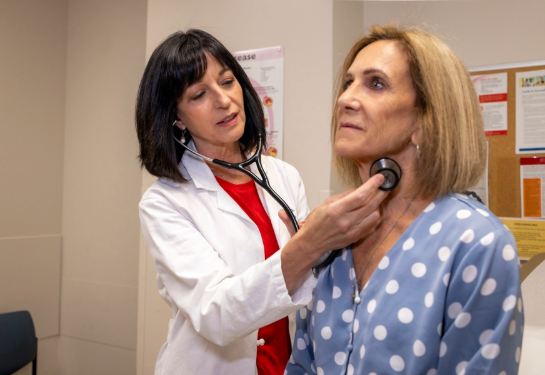 A person wearing a white coat uses a stethoscope to examine another person’s neck in a medical examination room with health posters visible in the bac