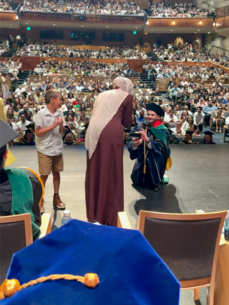 A man in blue graduation gown kneels before a woman standing on stage next to a boy in front of a large audience in auditorium