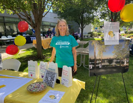 Blonde woman standing behind a table draped in yellow cloth and covered with brochures and other handouts.
