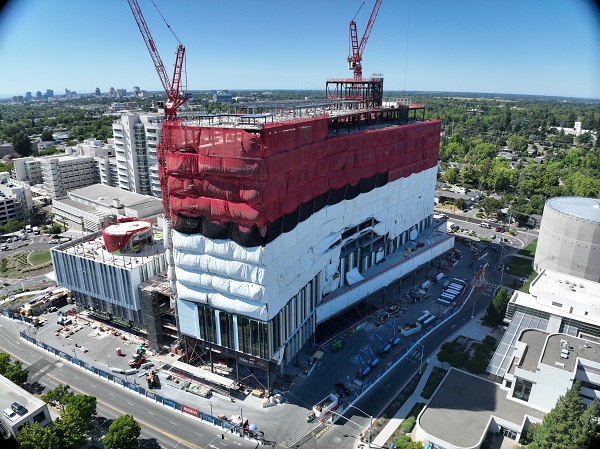Aerial view of California Tower under construction with red safety netting, white exterior wrap and two cranes above the structure on the Sacramento campus.