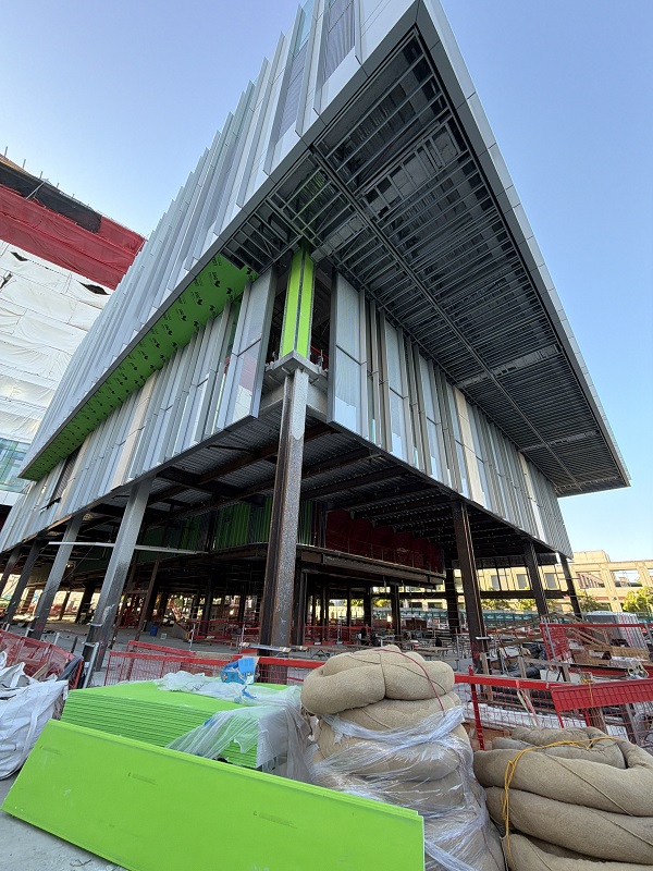 Low-angle exterior view of building fa&ccedil;ade panels and structural supports with construction materials in the foreground.