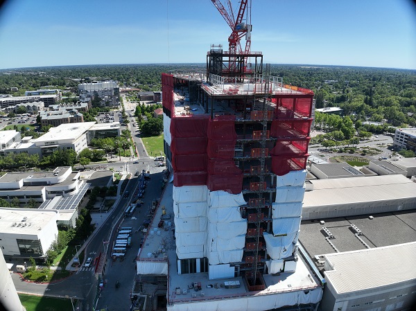 Aerial view of California Tower under construction with red safety netting, white exterior wrap and two cranes above the structure on the Sacramento campus.