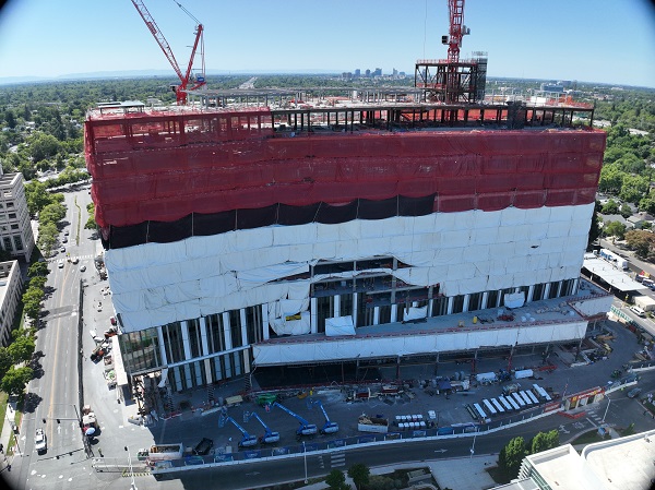 Aerial view of California Tower under construction with red safety netting, white exterior wrap and two cranes above the structure on the Sacramento campus.