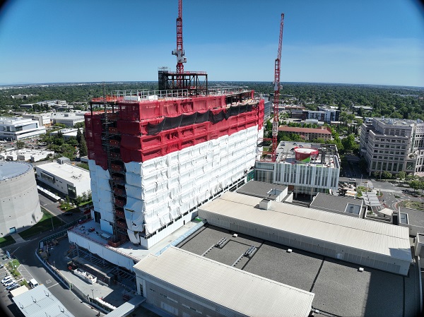 Aerial view of California Tower under construction with red safety netting, white exterior wrap and two cranes above the structure on the Sacramento campus.