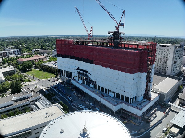 Aerial view of California Tower under construction with red safety netting, white exterior wrap and two cranes above the structure on the Sacramento campus.