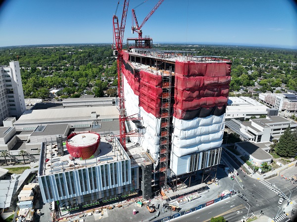 Aerial view of California Tower under construction with red safety netting, white exterior wrap and two cranes above the structure on the Sacramento campus.
