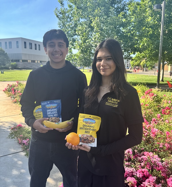Two individuals stand on a campus walkway holding packaged snack mixes, a banana, and an orange, with a UC Davis Health building and landscaping in the background.