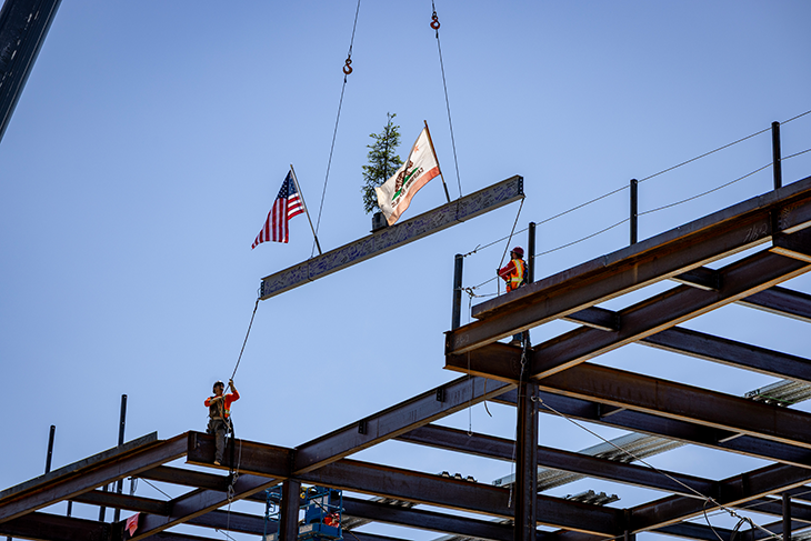 Construction workers guide a ceremonial steel beam into place during the topping out ceremony for the Central Utility Plant expansion project at UC Davis Health. The beam is decorated with an American flag, California state flag and evergreen tree.