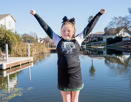 A young girl stands on a deck with water behind her. She wears a cheerleader uniform and stands with arms in a &ldquo;Y&rdquo; shape.