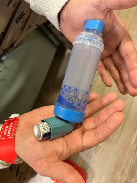 A close-up view of two hands holding a blue albuterol inhaler attached to a clear spacer device, with a red wristband.