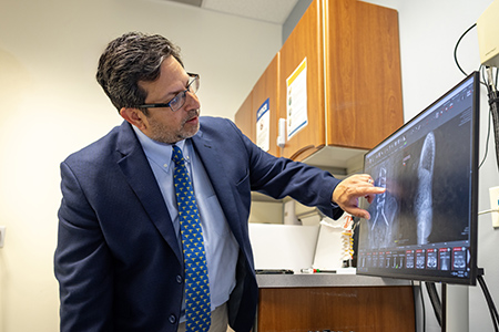 Man in dark blue suit points to two x-ray images on a computer screen. One image shows a close up of a spine with implants and screws. The other image is an opaque outline of a person&rsquo;s back.