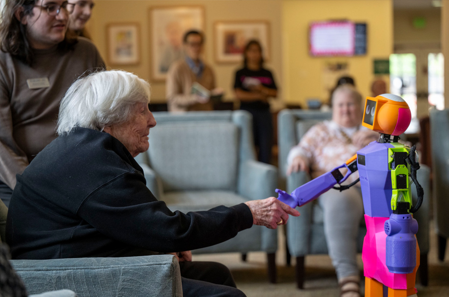 An older adult seated in a lounge reaches out to touch a colorful robot during an activity session.
