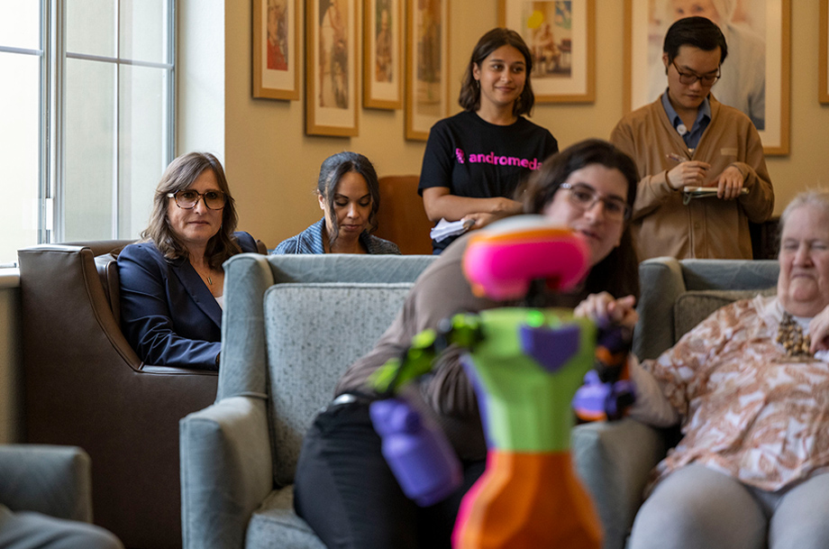 Researcher and others seated in a lounge watch as a caregiver demonstrates a colorful robot up close.