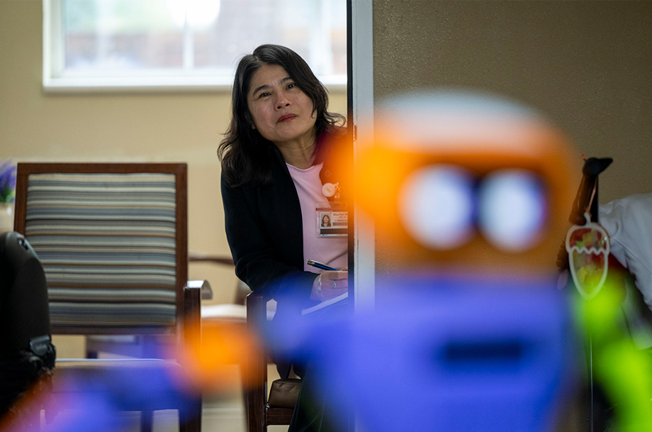 Researcher observes from a doorway while a colorful robot is in the foreground during an activity in a lounge.