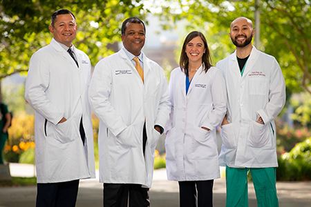 Three men and a woman standing in a row outside, all wearing white coats and posing with hands in pockets.