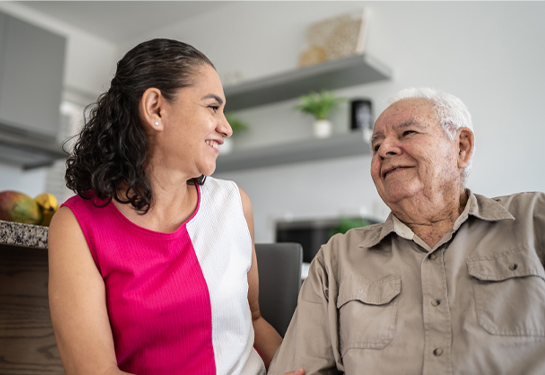 Woman smiling and talking with an older man while sitting together in a home setting.