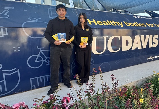 Two individuals stand near a UC Davis Health sign holding snack mixes and fruit, with flowers and campus buildings visible.