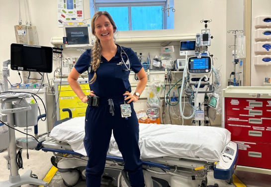A person wearing medical scrubs and a stethoscope stands in a hospital emergency room by a gurney, medical equipment and monitors.