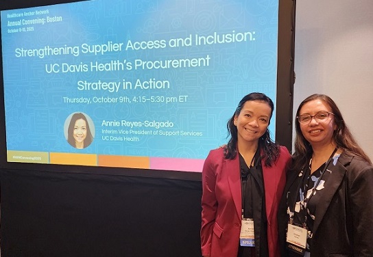 Two attendees stand in front of a presentation screen titled “Strengthening Supplier Access and Inclusion: UC Davis Health’s Procurement Strategy