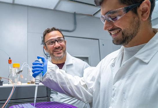 Two researchers in white coats and lab goggles work in a lab.