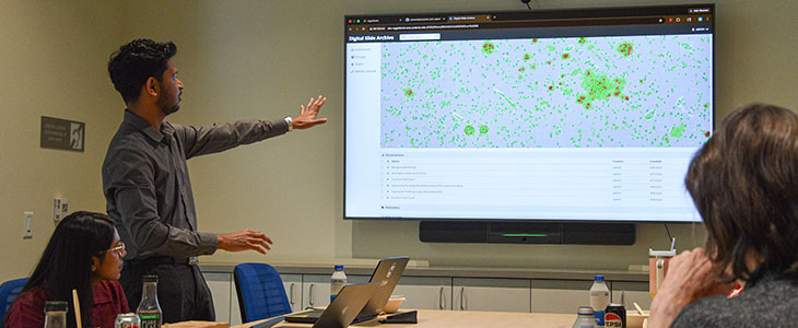 A man stands near a whiteboard, pointing to a slide that shows cells from a brain sample while two people look on. 