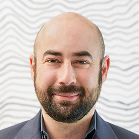 David Brandman, in a dark blue shirt, and with a beard, stands before a while wall, smiling for the camera.
