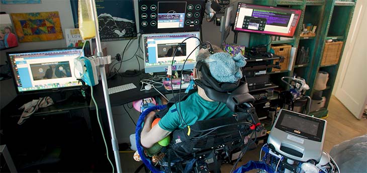 A man in a wheelchair sits behind a desk set up with a brain computer interface and multiple monitors. 