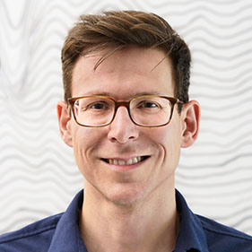 Sergey Stavisky, in glasses and a navy blue shirt, stands before a white wall, smiling at the camera.