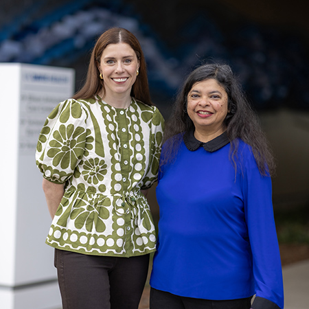 A woman with light brown hair, wearing a bright green print blouse, standing next to dark haired woman with royal blue blouse.