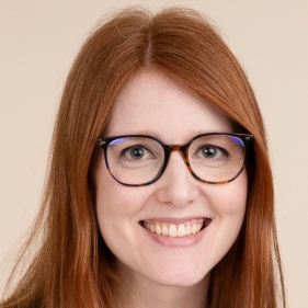 Smiling person with long red hair and glasses facing the camera against a plain beige background.