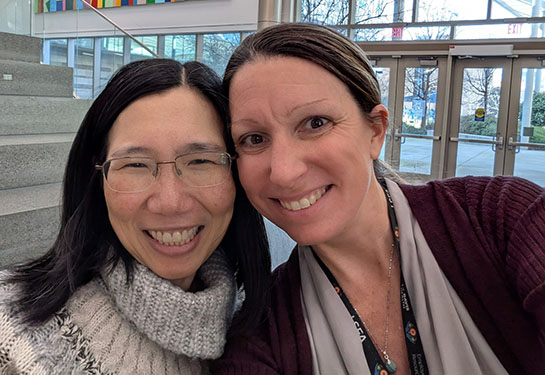 Two women stand inside a brightly lit building, faces close together and smiling to the camera. 