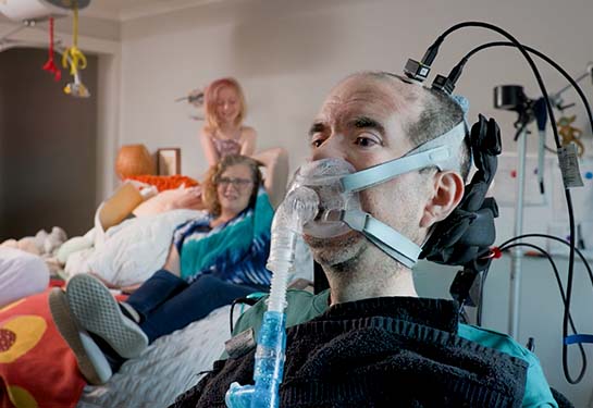 A man using a breathing apparatus and a brain-computer interface sits in a chair in the foreground while his wife and daughter sit on a bed behind him