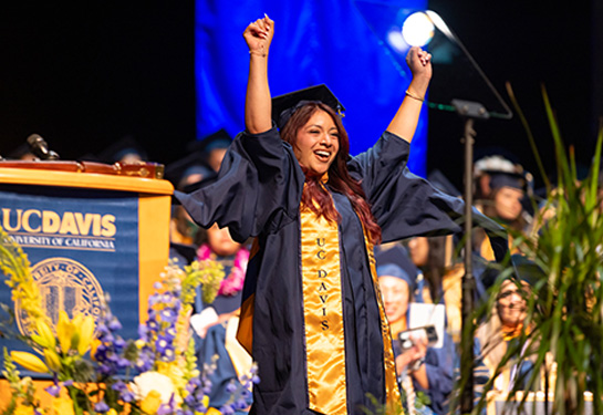 Graduate in cap and gown raises both arms in celebration on stage during commencement, smiling as classmates and faculty look on.